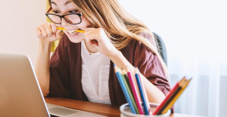 A woman is biting a pencil while looking at the content on her laptop